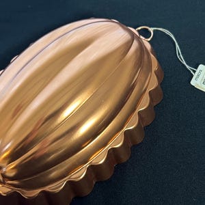 May include: A copper-colored, oval-shaped mold with a scalloped edge. The mold has a ribbed design on the top surface and a small ring for hanging. A tag attached to the mold reads "macys".