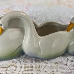 May include: A ceramic swan planter with three swan figures. The planter is primarily white with yellow beaks and subtle blue and green accents. The swan's necks curve to form the planter's openings.