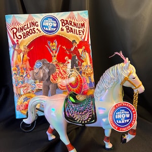 May include: A pearlescent white circus horse figurine with a saddle and decorative accents. The horse has red and orange leg wraps and a tag that reads "The Greatest Show on Earth." A Ringling Bros. and Barnum & Bailey circus poster is in the background.