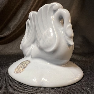 May include: A light blue ceramic swan figurine with a raised base. The swan has a detailed design and a glossy finish. The base has a label that reads "Hallmark China".
