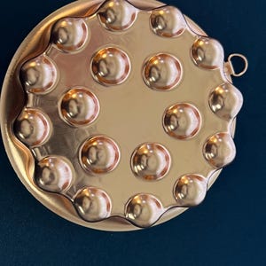 May include: A gold-colored escargot plate with 12 circular indentations. The plate has a scalloped edge and a small handle. The surface of the plate reflects light, creating highlights and shadows. The plate is designed for serving escargot.