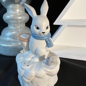 May include: A white porcelain figurine of a rabbit wearing a blue scarf and holding a fishing net. The rabbit is standing on a white porcelain base that resembles a snowdrift. A blue fish is partially submerged in the snowdrift.