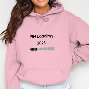 Peut inclure: Un sweat à capuche rose clair avec le texte "RN Loading ... 2026" en noir. En dessous, un graphique de barre de chargement. Le sweat à capuche a une capuche à cordon et une poche avant.
