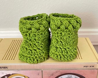 May include: A pair of handmade, green crochet baby booties. The booties are a vibrant green color and have a textured, bobble stitch design. They are displayed on a vintage radio.