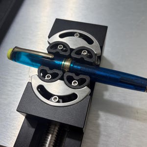May include: A blue fountain pen with gold detailing is held in a black and silver pen vice. The pen has a yellow tip and a transparent blue barrel. The vice has a screw mechanism and is designed to hold pens securely.