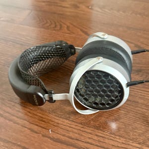 May include: Silver and black over-ear headphones with a honeycomb design on the earcups. The headphones have a black headband and ear pads, and a silver frame. The headphones are on a wooden surface.