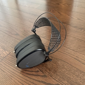 Puede incluir: Auriculares negros de diadema con una diadema negra y un diseño de malla negra. Los auriculares están sobre una superficie de madera.