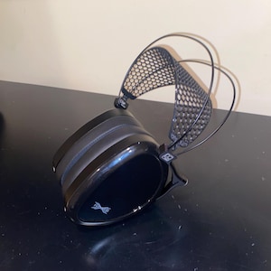 Puede incluir: Auriculares negros con diadema de malla negra y un logotipo negro y plateado en la almohadilla.