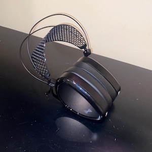 Puede incluir: Auriculares negros de diadema con diadema de fibra de carbono y almohadilla de malla negra. Los auriculares est&aacute;n sobre una superficie negra.