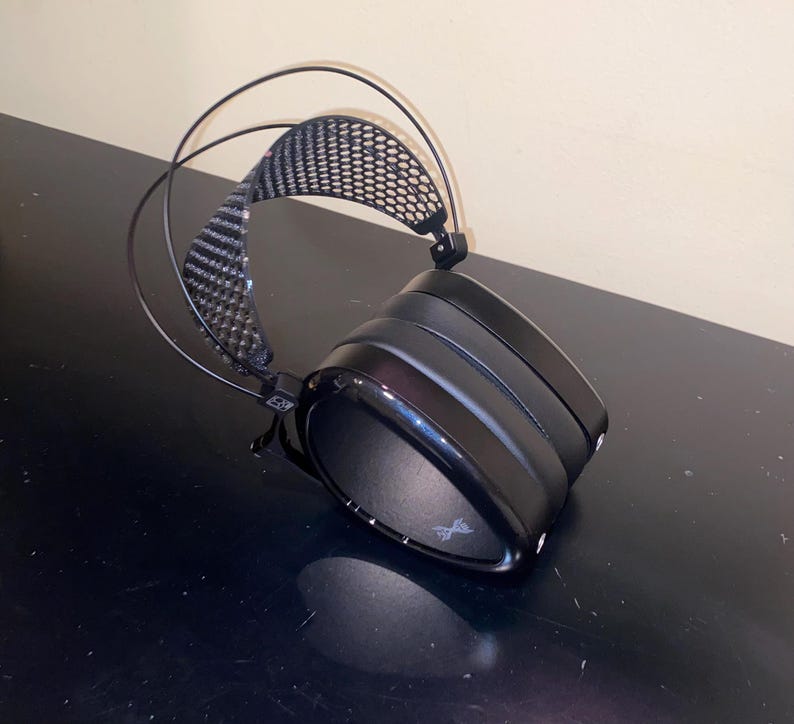 Puede incluir: Auriculares negros de diadema con diadema de fibra de carbono y almohadilla de malla negra. Los auriculares est&aacute;n sobre una superficie negra.