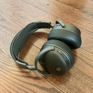 May include: Black over-ear headphones with a black headband and a mesh design. The headphones have a volume control and a button on the side.