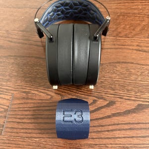 May include: Black headphones with a blue and black padded headband. The headphones are sitting on a wooden surface. A blue cylindrical object with the text "E3" is in the foreground.