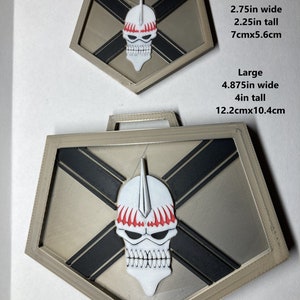 Puede incluir: Dos medallones de metal de color plateado con un diseño de calavera. El medallón más pequeño mide 2,75 pulgadas de ancho y 2,25 pulgadas de alto. El medallón más grande mide 4,875 pulgadas de ancho y 4 pulgadas de alto. Ambos medallones tienen un diseño de cruz en blanco y negro con una calavera blanca en el centro. La calavera tiene acentos rojos alrededor de los ojos y en la parte superior de la cabeza.