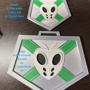 Puede incluir: Dos medallas hexagonales plateadas con un diseño verde y blanco. La medalla más grande mide 12,2 cm de ancho y 10,4 cm de alto. La medalla más pequeña mide 7 cm de ancho y 5,6 cm de alto.