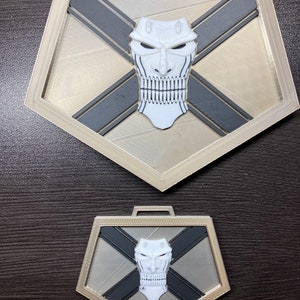 May include: Two 3D printed pendants, one larger than the other, both featuring a white skull with a cross-hatch pattern on a gold background. The skull has a menacing expression with a toothy grin.