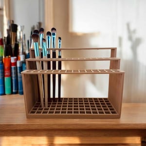Puede incluir: Un soporte para pinceles de madera de tres niveles con un patrón de rejilla. El soporte está hecho de madera marrón claro y tiene un patrón de rejilla en cada nivel. Hay varios pinceles en el soporte, algunos con mangos azules y negros.