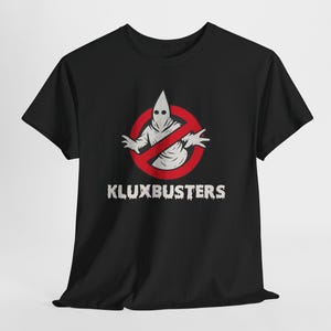 May include: Black t-shirt featuring a parody design. The graphic includes a hooded figure inside a red no-entry circle, with the text "KLUXBUSTERS" in a distressed font. The design is centered on the front of the shirt.