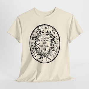 May include: A cream-coloured t-shirt with a black oval design featuring text and laurel leaves. The text reads "SOCIETE DES AMIS DE LA CONSTITUTION" and "VIVRE LIBRE OU MOURIR."