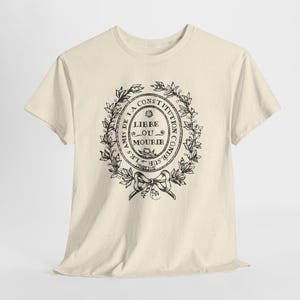 May include: Cream-colored t-shirt featuring a black circular design with text and a floral wreath. The text reads "LES AMIS DE LA CONSTITUTION CONDÉ SUR NOTHAIN LIBRE OU MOURIR". A bow is at the bottom of the design.
