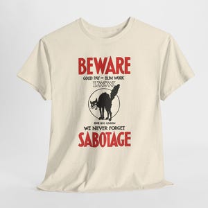 May include: A cream-colored short-sleeve t-shirt featuring a graphic print with the red text BEWARE and SABOTAGE. The design includes a black illustration of a hissing cat inside a circle, with the text GOOD PAY OR BUM WORK, I.W.W., and ONE BIG UNION WE NEVER FORGET.