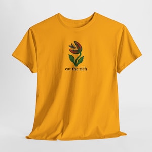 May include: Mustard yellow t-shirt featuring a graphic of a Venus flytrap with a red and green flower and the text "eat the rich" in black. The shirt is made of a soft material.