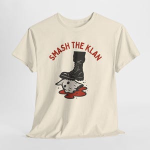 May include: Cream-coloured t-shirt with the red text "SMASH THE KLAN" above a graphic of a black boot stepping on a ghost-like figure with red blood. The shirt is made of a soft material.