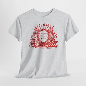 May include: A pale grey t-shirt with a red graphic. The design features the text "GUERRE AUX ENNEMIS DU PEUPLE" within a decorative emblem of flags, swords, and other elements. The t-shirt has short sleeves and a crew neck.