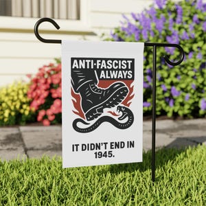 May include: A white garden flag with a black metal stand. The flag features the text "ANTI-FASCIST ALWAYS" above an illustration of a boot stepping on a snake. Below the illustration is the text "IT DIDN'T END IN 1945."