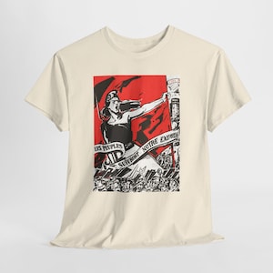 May include: A cream-coloured short-sleeve t-shirt featuring a graphic print in red, black, and white. The design depicts a figure holding a large red flag with the text LES PEUPLES SUIVRONT NOTRE EXEMPLE above a crowd of people with raised tools.