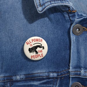 May include: A circular white badge with the words "ALL POWER TO THE PEOPLE" in red and black, and a black panther illustration. The badge is attached to a blue denim jacket.