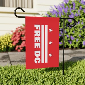 May include: A red garden flag with the text "FREE DC" in white, along with three white stars and two vertical white stripes. The flag is displayed on a black metal stand in a grassy yard, with colorful flowers in the background.