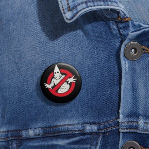 May include: A black button pin with a white figure in a pointed hood and a red prohibition sign. The pin is set against a blue denim jacket.
