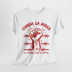 May include: White t-shirt with a red graphic. The graphic features a clenched fist within barbed wire, with the words "CHINGA LA MIGRA" above and "NO HUMAN BEING IS ILLEGAL" below. The design is centered on the shirt.