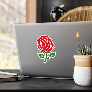 May include: A die-cut sticker featuring a stylised red rose graphic with the letters DSA integrated into the petals. The design includes a green stem and leaves, outlined in white, applied to the centre of a silver laptop lid surface.