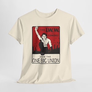 May include: A cream-colored short-sleeve t-shirt featuring a graphic print with a red background. The design shows a person with one arm raised, industrial silhouettes, and the text: THE I.W.W. IS COMING! JOIN THE ONE BIG UNION.