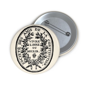 May include: Cream-colored button with a black graphic design and text. The design features a wreath surrounding the words "Vivre Libre Ou Mourir" and other text. A second button is visible in the background.