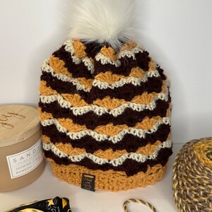May include: A crocheted beanie hat with a brown, white, and mustard yellow chevron pattern. The hat has a white pom pom on top and a brown leather tag that says "Made with Love by Erin".