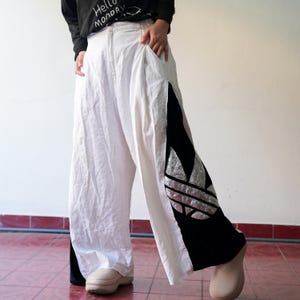 May include: White wide-leg pants with a black and silver Adidas logo on the side. The pants have a relaxed fit and a high waist.