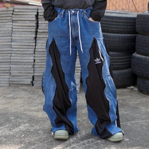 Puede incluir: Pantalones vaqueros azules con paneles laterales negros ondulados y cordón blanco. Los pantalones presentan el logo de Adidas en el panel negro. Los pantalones tienen un corte holgado y se llevan con zapatos verdes.