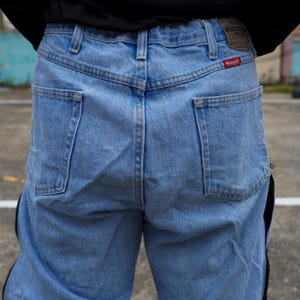 May include: Light blue denim jeans with the Wrangler logo on the back pocket. The jeans have a classic five-pocket design and a straight leg cut. The back of the jeans are visible, showing the pockets and the waistband.