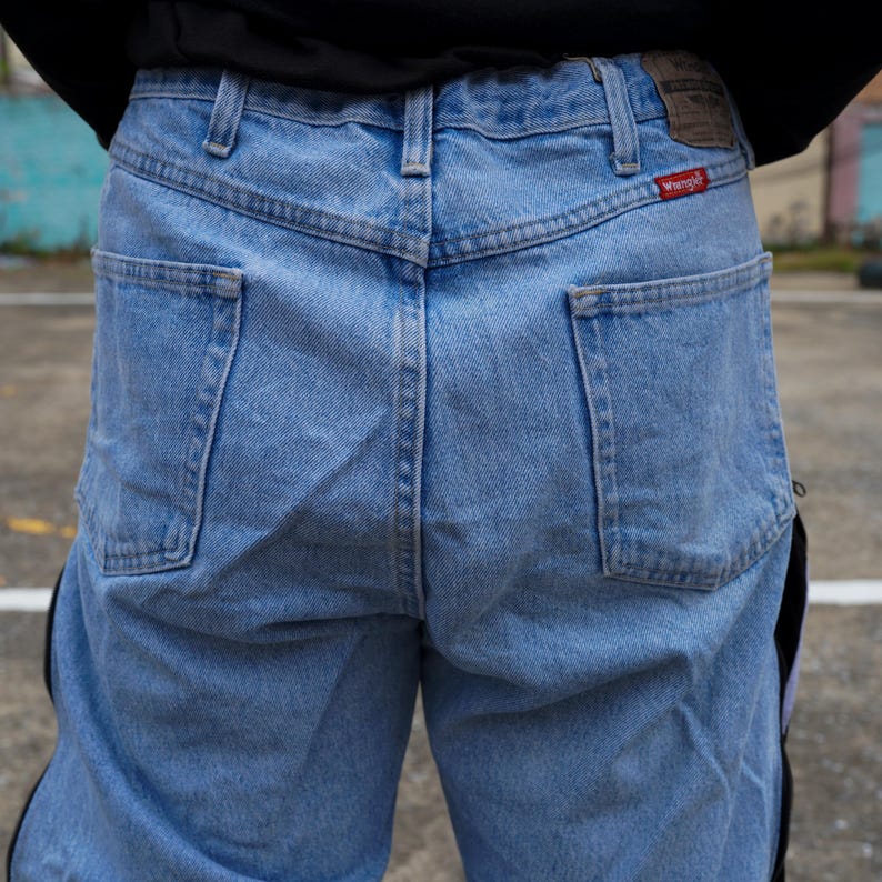 May include: Light blue denim jeans with the Wrangler logo on the back pocket. The jeans have a classic five-pocket design and a straight leg cut. The back of the jeans are visible, showing the pockets and the waistband.