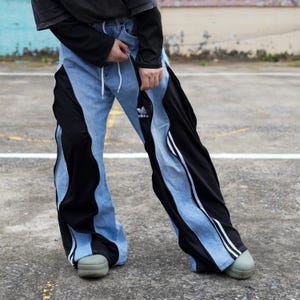 May include: Light blue denim pants with black side panels and white stripes. The pants feature a drawstring waist and the Adidas logo. The pants have a unique, modern design.