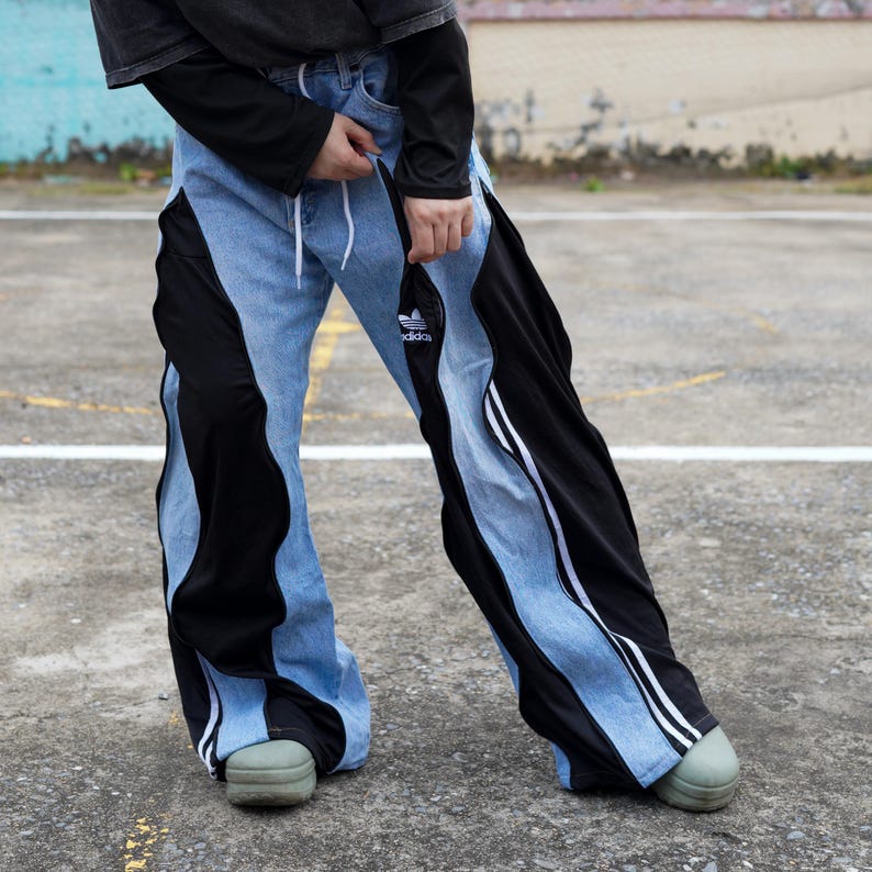 May include: Light blue denim pants with black side panels and white stripes. The pants feature a drawstring waist and the Adidas logo. The pants have a unique, modern design.