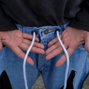May include: Close-up of light blue denim jeans with a drawstring. The jeans have a button closure and black accents. White drawstrings extend from the waistband. The person is wearing a dark gray long-sleeve shirt.