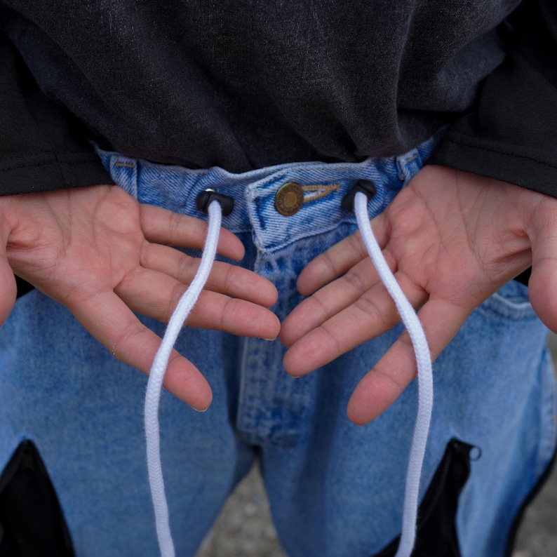 May include: Close-up of light blue denim jeans with a drawstring. The jeans have a button closure and black accents. White drawstrings extend from the waistband. The person is wearing a dark gray long-sleeve shirt.