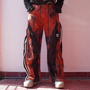 Peut inclure: Pantalon large avec un motif tie-dye dans des tons orange et noir. Le pantalon présente des panneaux latéraux noirs avec des bandes blanches et le logo Adidas sur la cuisse gauche. Il possède une taille à cordon et une coupe ample et décontractée.