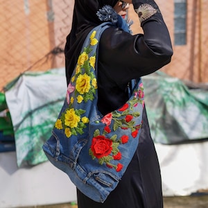 Może przedstawiać: Dżinsowa torba na ramię z żywą haftowaną kwiatami. Torba ma żółte, czerwone i różowe kwiaty, tworzące kolorowy kontrast z niebieskim dżinsem. Unikalny, ręcznie robiony dodatek.