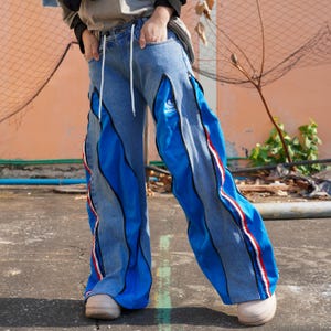 May include: Blue denim jeans with blue and white striped panels down the sides. The jeans have a white drawstring and the word "adidas" is visible on the right leg.