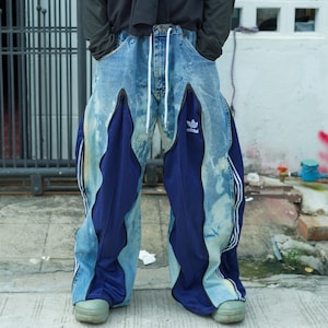 Op de afbeelding: Blauwe spijkerbroek met donkerblauwe panelen en witte strepen. De broek heeft een taille met trekkoord en een Adidas-logo. Het ontwerp is uniek, asymmetrisch en heeft een vervaagd waseffect.