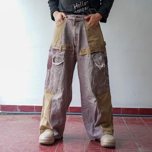 Op de afbeelding: Wijd uitlopende patchwork jeans in bruin- en mauve tinten. De jeans heeft grote zakken, riemlussen en contrasterende panelen. De persoon draagt een zwart shirt met lange mouwen met de tekst "Hello Monday".